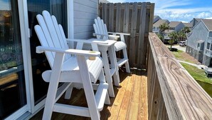 Un terrazzo in legno con poltrone Adirondack bianche e un tavolo, con vista su una zona residenziale con case e cielo sereno.