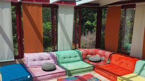A sunlit outdoor seating area with cushioned sofas and ottomans in various colors, surrounded by large windows and a view of greenery. - Magnificent villa in the heart of the Var for a superb vacation. (La Verdiere)