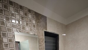 Baño con lavamanos blanco, inodoro y espejo. Hay un estante con artículos de aseo y una ventana sobre el inodoro.