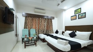 Habitación de hotel con una cama grande, una zona de estar con dos sillones, una mesita con un jarrón y un televisor montado en la pared.