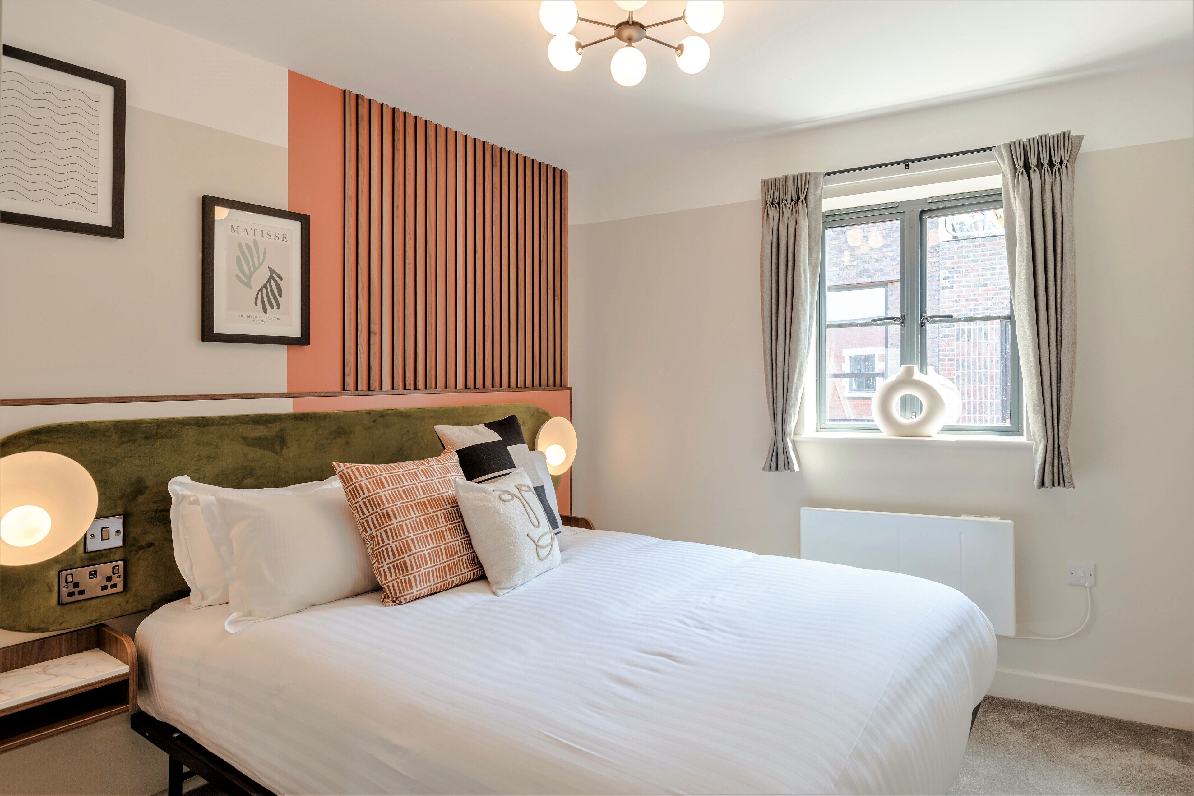 Une chambre à coucher bien rangée, avec un lit, des lampes de chevet, une œuvre d’art encadrée et une fenêtre avec des rideaux.
