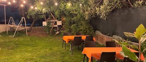 Un área de comedor al aire libre con mesas, sillas, luces colgantes y plantas.