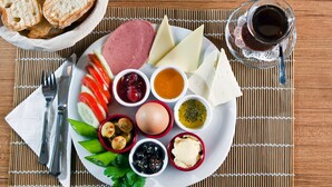 A plate with various cheeses, meats, and condiments, accompanied by bread and a glass of drink on a bamboo mat.