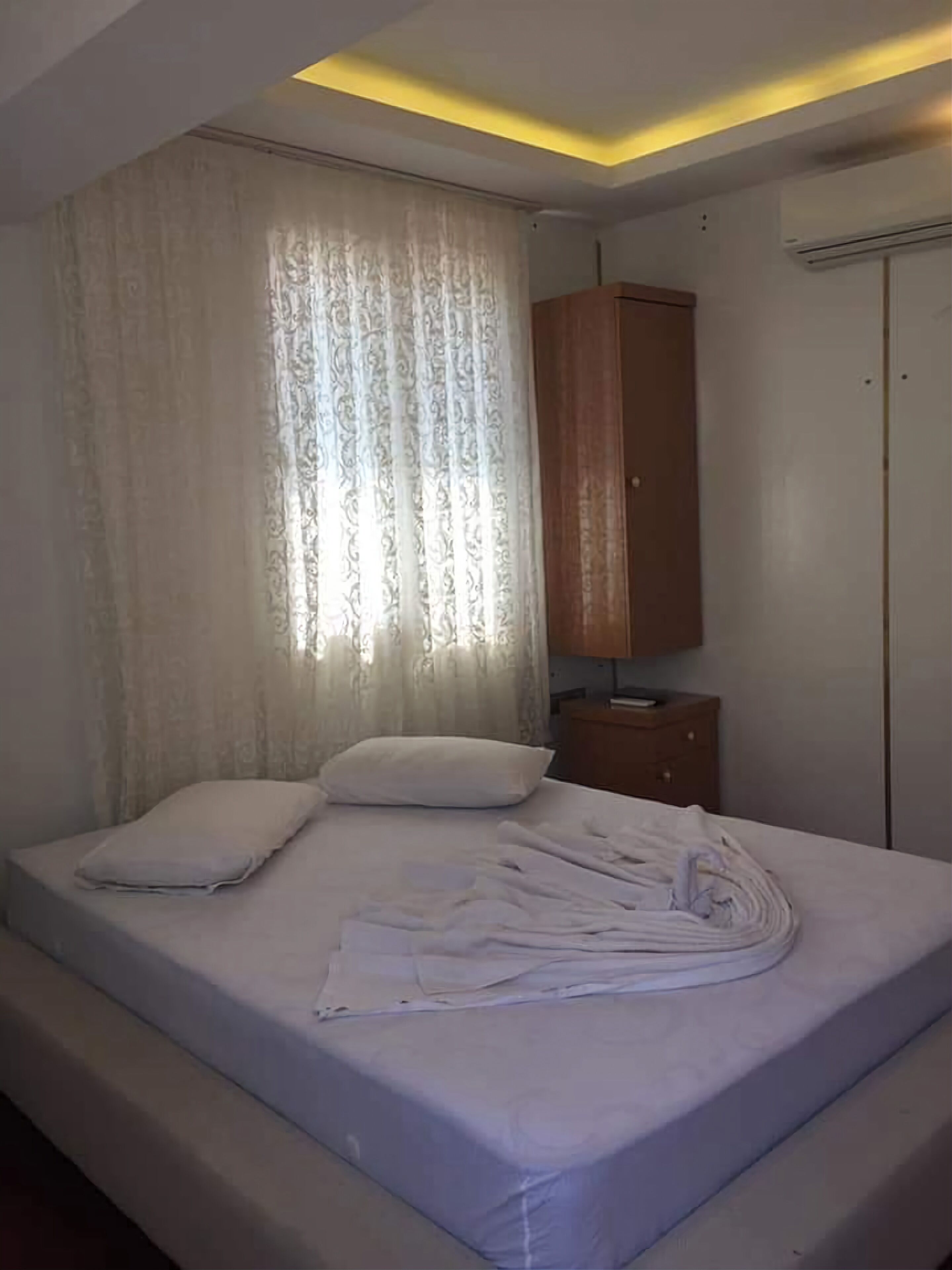 A neatly made bed with pillows, a wooden nightstand, a wardrobe, and a window with curtains.