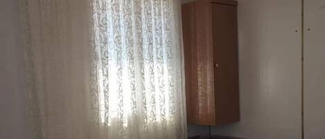 A neatly made bed with pillows, a wooden nightstand, a wardrobe, and a window with curtains.
