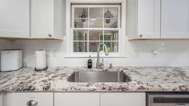 A kitchen with white cabinets, a double sink, a marble countertop, and a window with plants. - Pet-Friendly 3BR 2BA - Fenced Yard (Durham)