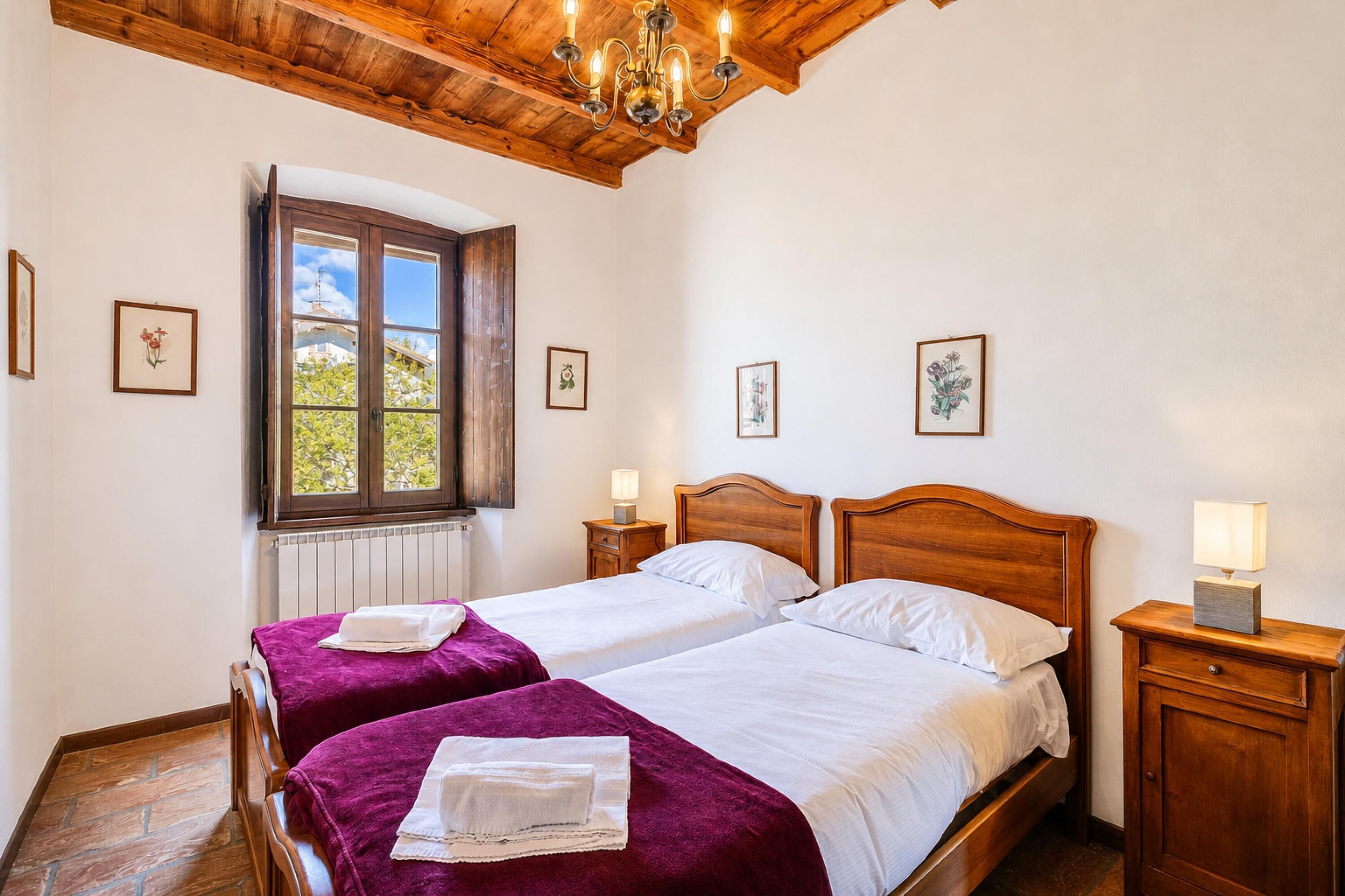 A room with two beds, wooden headboards, a wooden ceiling, a window with a view, and framed pictures on the walls.