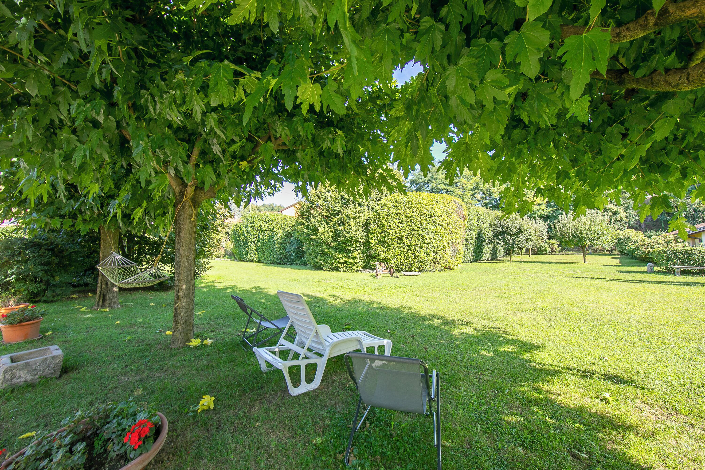 Ein grünes Areal im Freien mit zwei weißen Liegestühlen unter einem Baum, einer Hängematte und einem kleinen Garten mit roten Blüten.