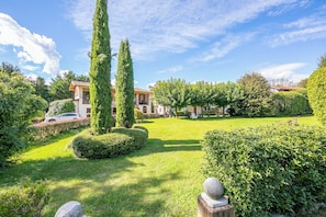 Um jardim bem cuidado, com sebes, um banco de pedra e uma árvore alta.