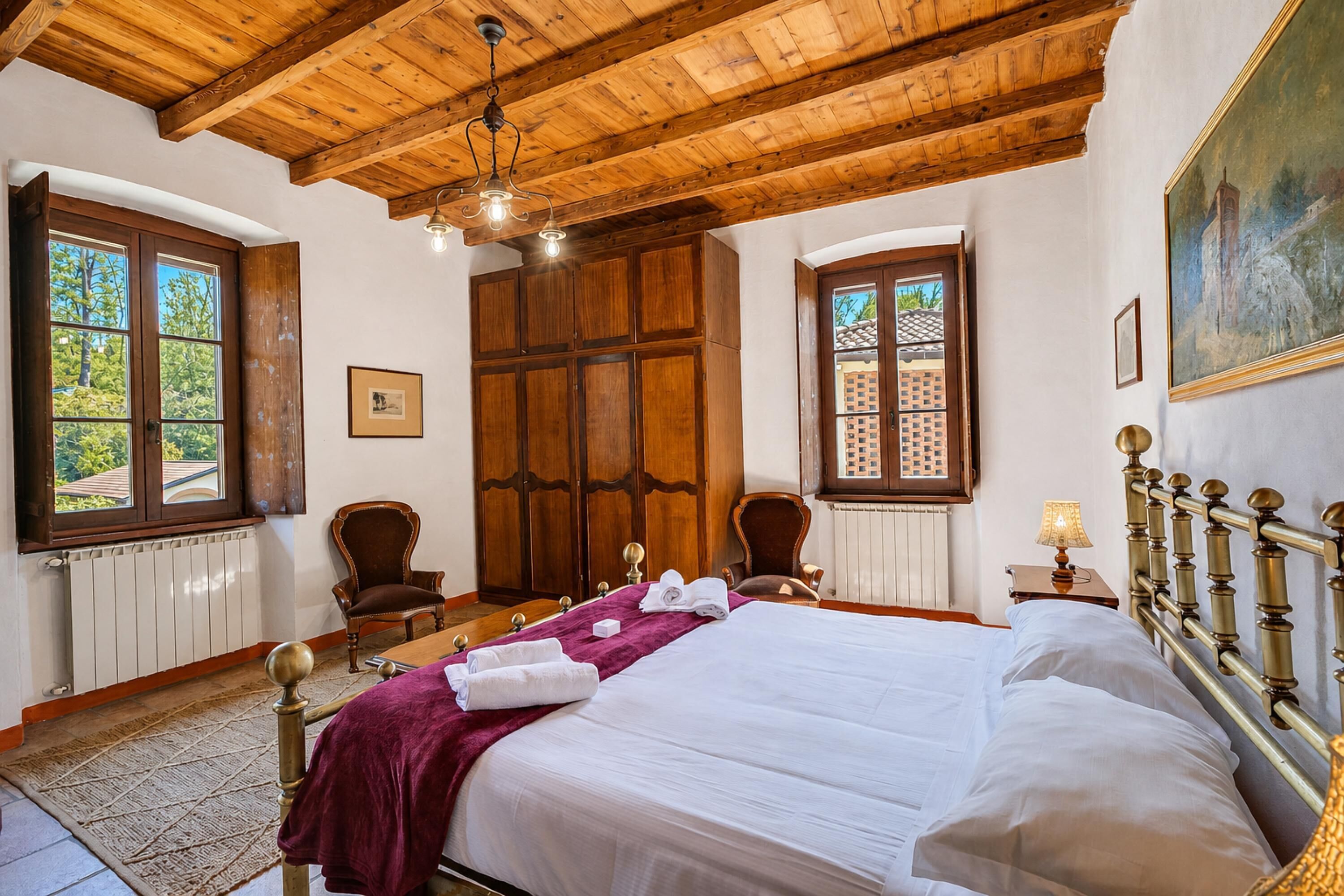 Un dormitorio con techo de madera, una cama grande, dos sillas, un armario de madera y una ventana con vistas a árboles.