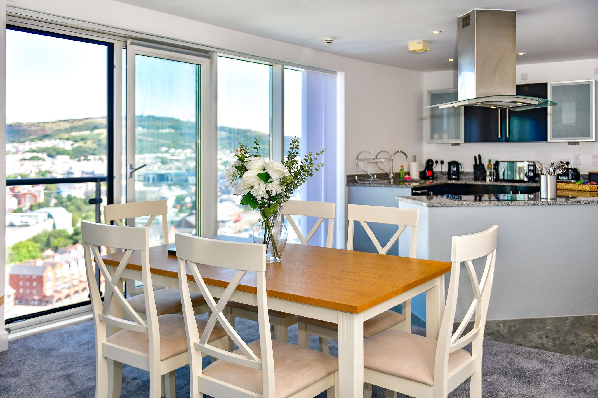 A dining area with a wooden table and white chairs, a vase with flowers, and a city view through large windows.