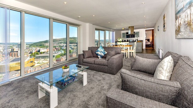 A spacious living area with a large window offering a city view, a modern kitchen in the background, and a comfortable seating arrangement. - Just Stay Wales - Meridian Tower (Swansea)