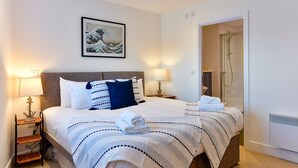 A neatly made bed with striped bedding, a framed artwork above it, and a visible shower area to the right. - Just Stay Wales - Meridian Tower (Swansea)