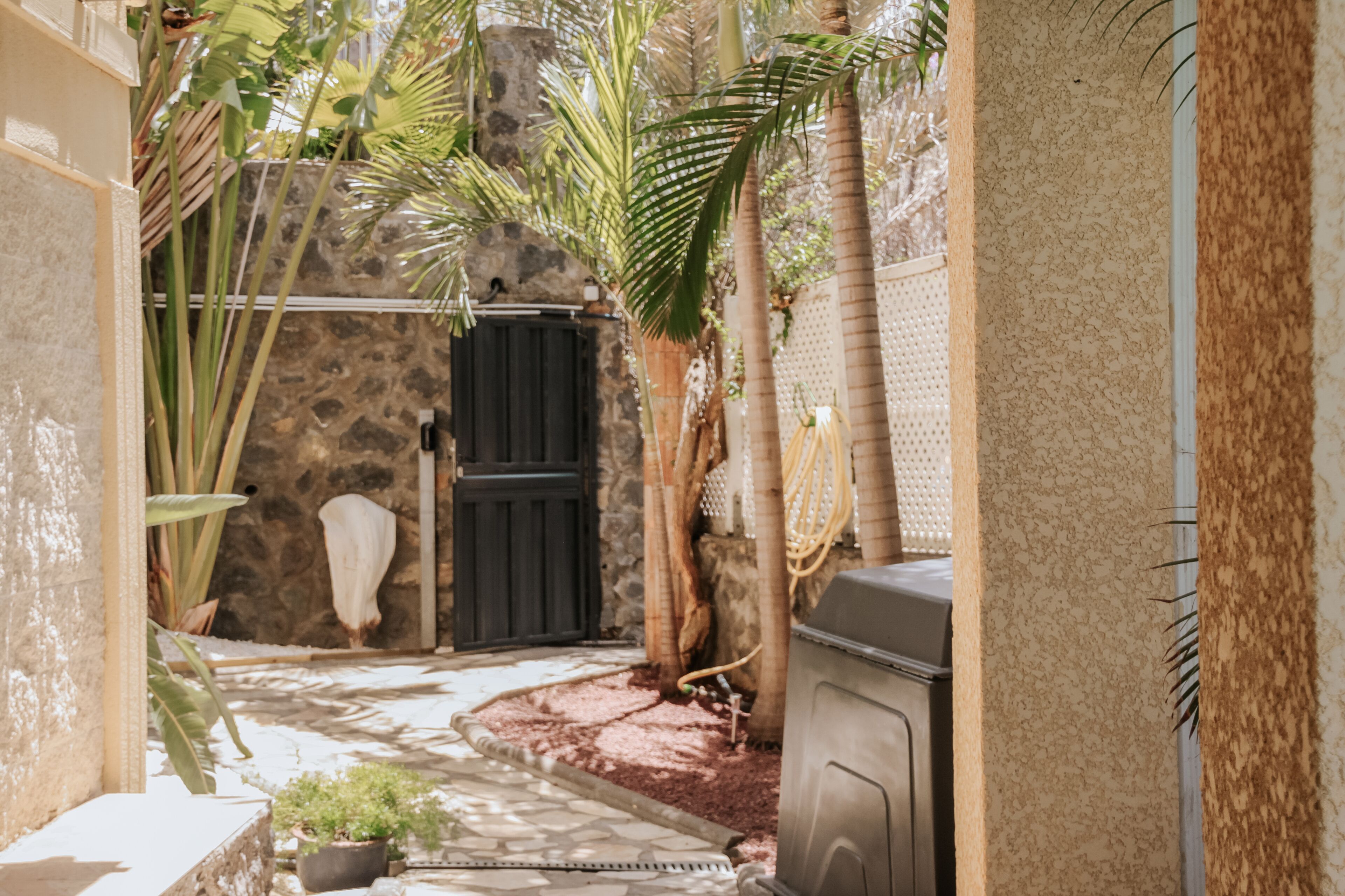 A narrow outdoor pathway leading to a stone wall with a black gate, surrounded by potted plants and a small garden area.
