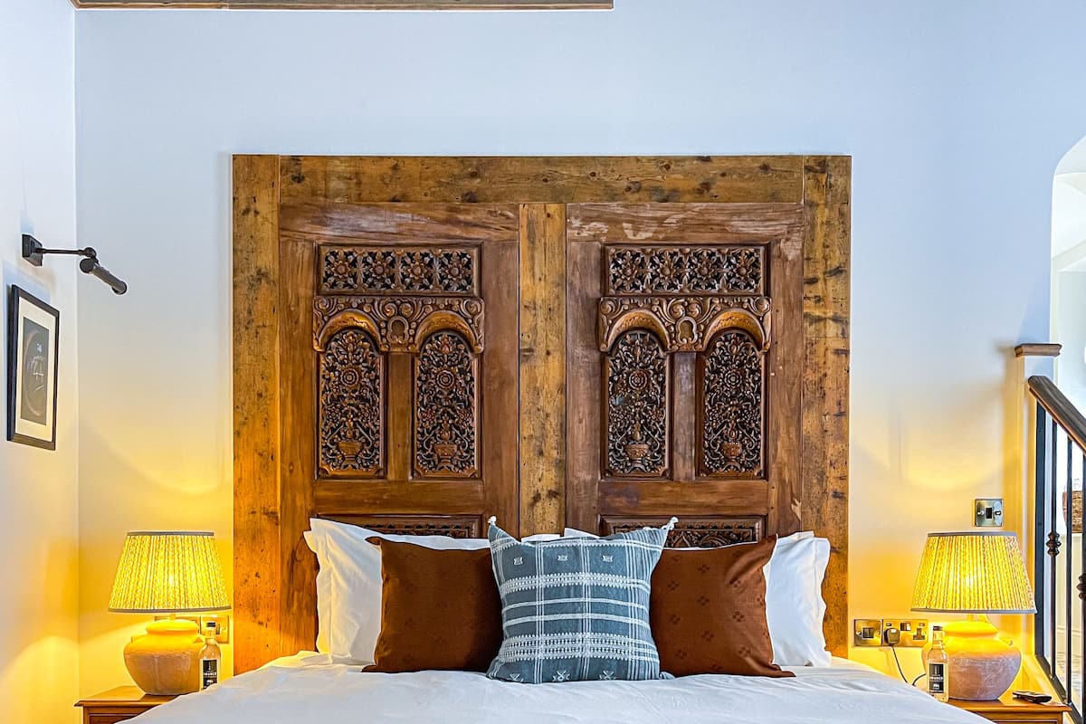 Un dormitorio con una cama grande, cabecera de madera, dos mesitas de noche con lámparas y una pequeña mesa de madera frente a la cama.