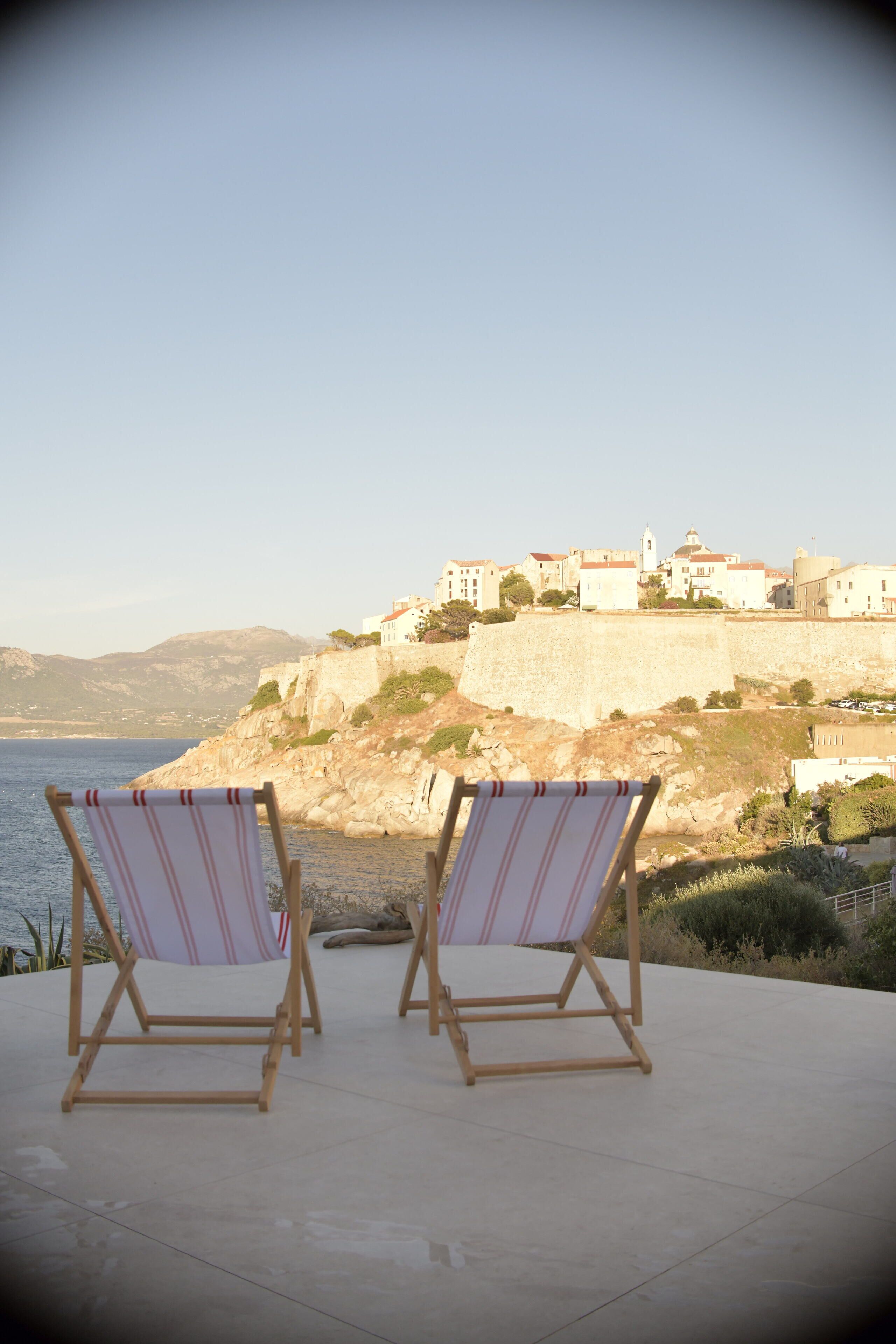 Dos sillones de madera con tela a rayas mirando hacia un pueblo costero con edificios históricos y vista al mar.