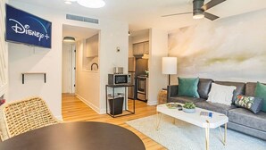 Ein modernes Wohnzimmer mit einem Flachbildfernseher, der das Disney+-Logo zeigt, einem grauen Sofa mit grünen und weißen Kissen, einem weißen Couchtisch und einem Deckenventilator.