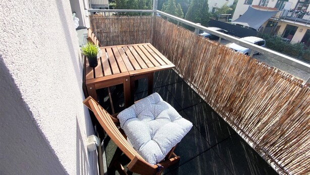 Un balcone con tavolo in legno, una sedia imbottita, una recinzione in bambù e vista su edifici e alberi.