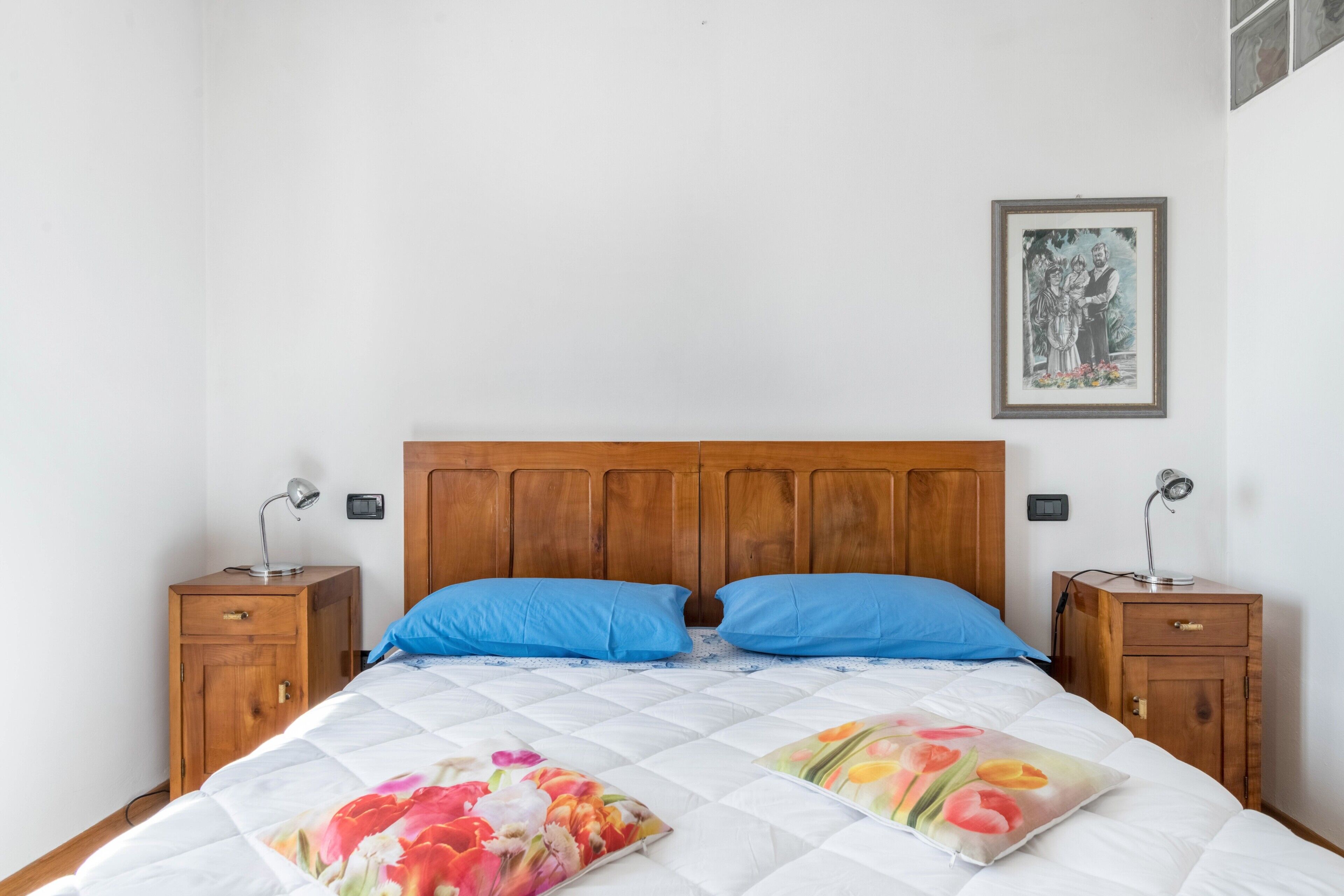 Una cama con cabecera de madera, dos almohadas azules, una colcha con estampado floral y una mesita de noche con lámpara.