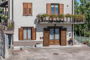 En toetagers bygning med træpersienner, en balkon med blomster og et lille udendørs siddeområde.