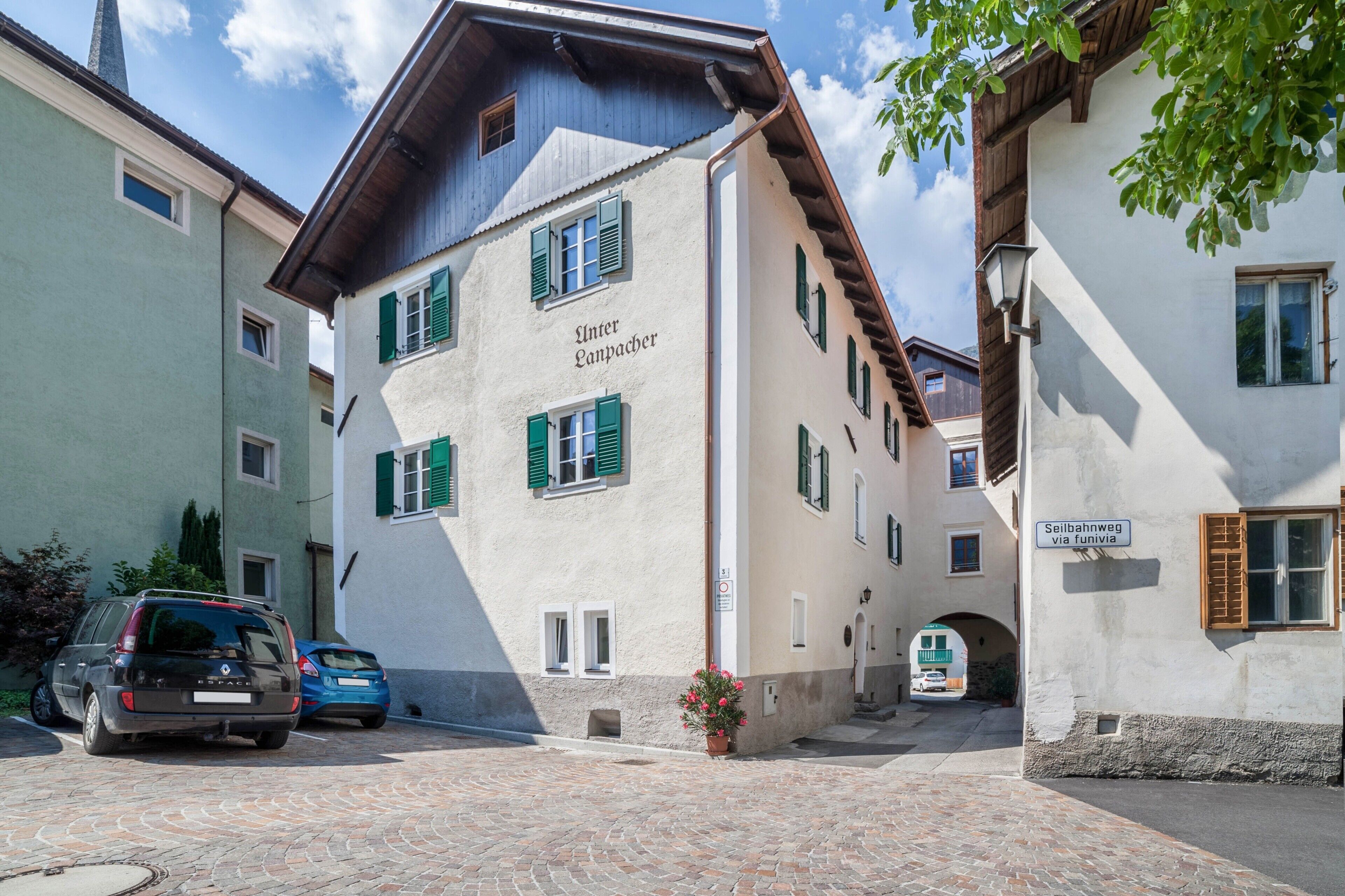 A building with a sign reading 'Unter Langpacher' and another sign 'Seilbahnweg via Funivia.' There are parked cars and a cobblestone street.