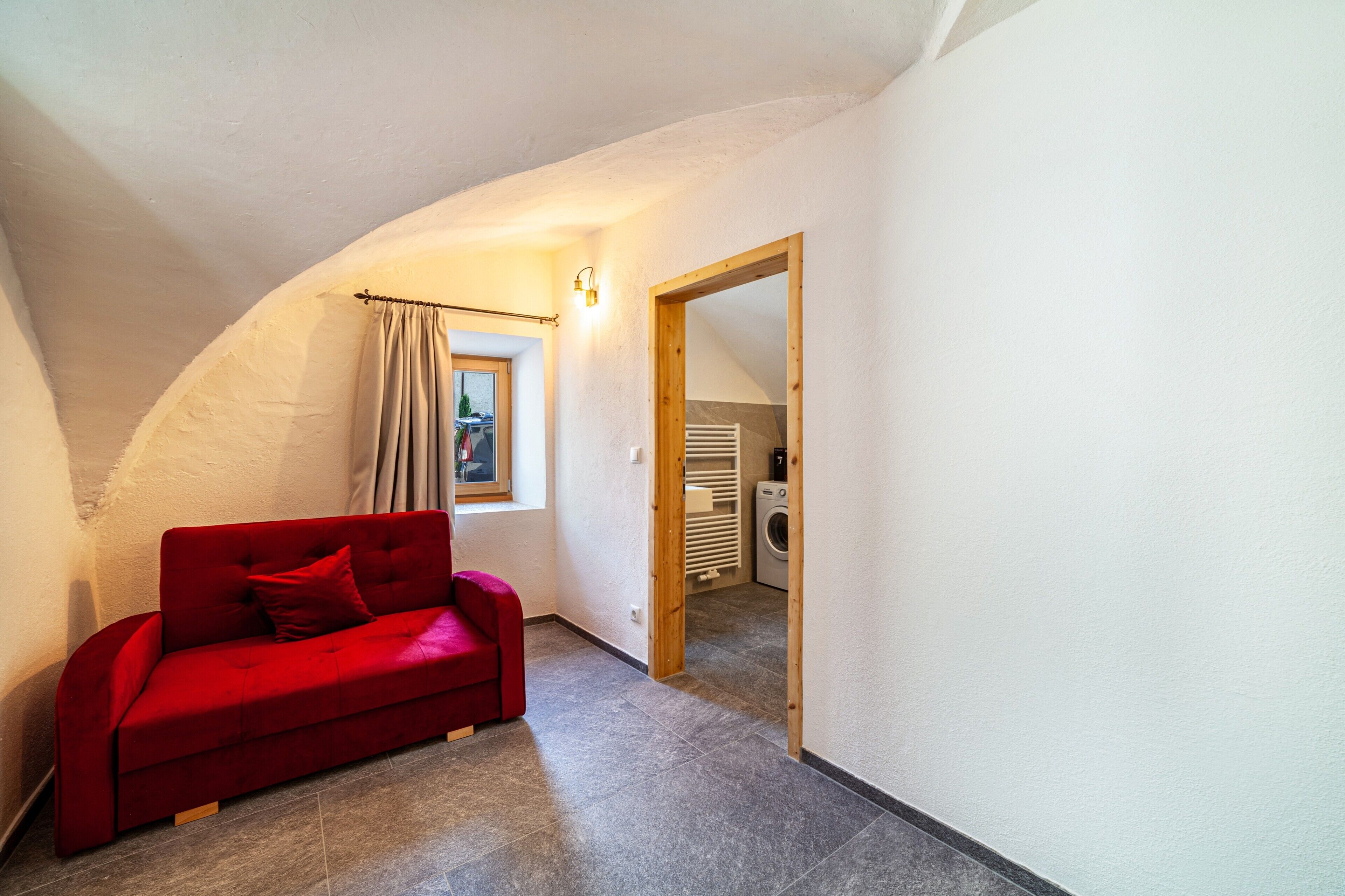 A compact room with a red sofa, a washing machine, and a small window with curtains.