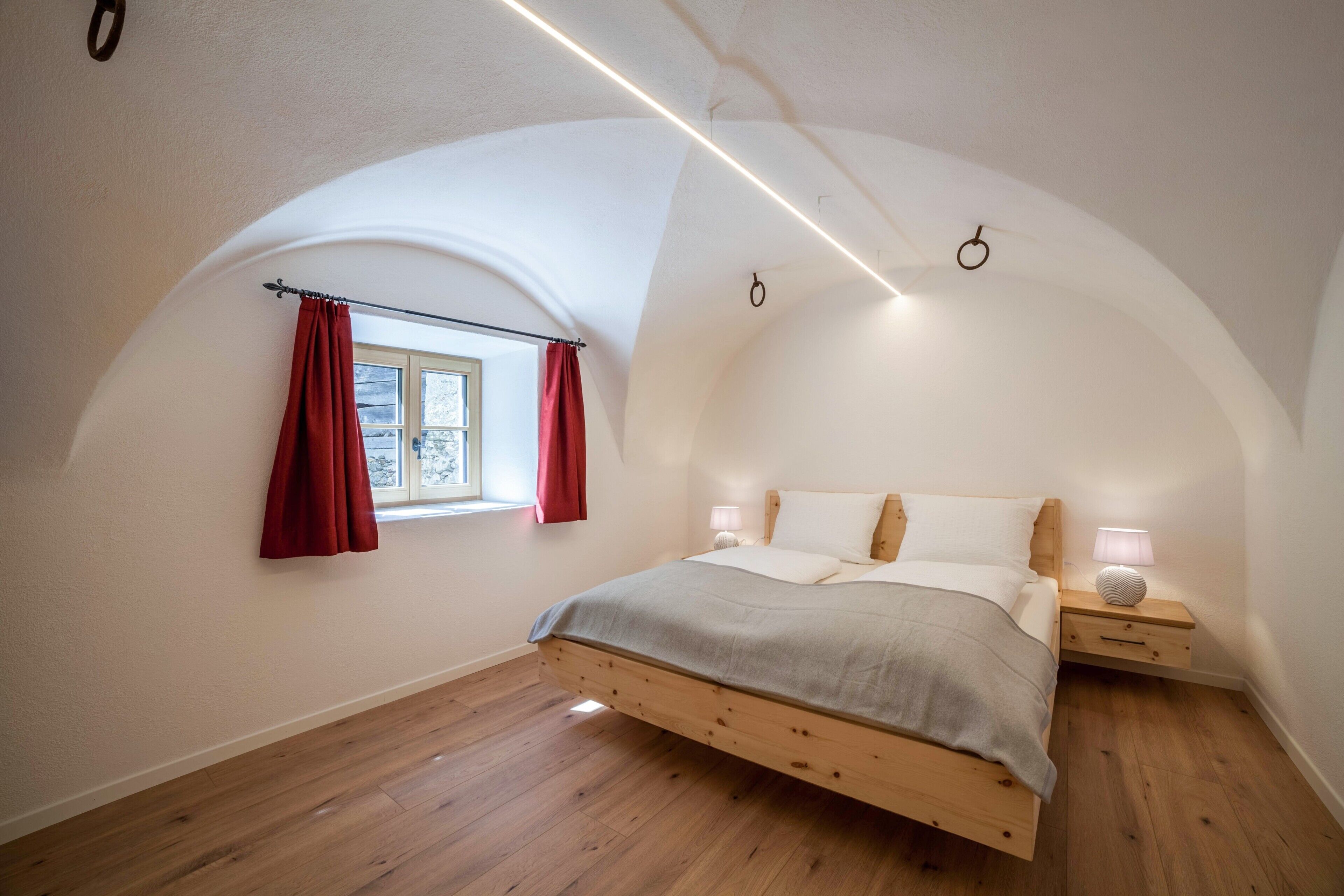 Een slaapkamer met een schuine plafond, een bed met houten frame, een nachtkastje met lamp en een raam met gordijnen.
