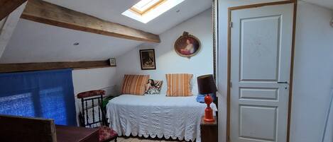 Un dormitorio con techo inclinado, vigas de madera, cama con ropa de cama estampada, una mesita con lámpara y una puerta con diseño circular.