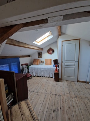 Une chambre avec un plafond en pente, des poutres en bois, un lit avec une literie à motifs, une petite table avec une lampe et une porte ornée d’un motif circulaire.