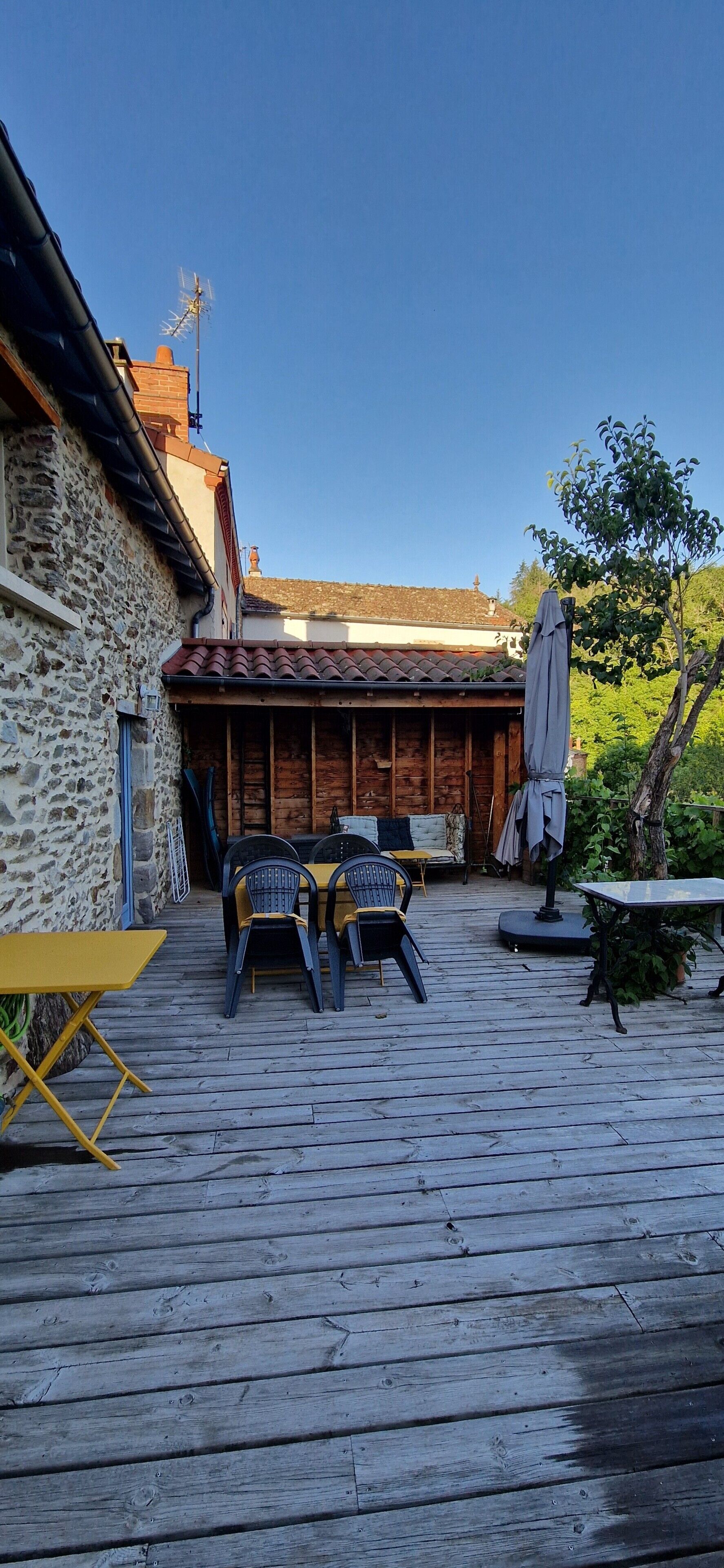 Una terraza de madera con mobiliario exterior, un muro de piedra y una estructura de techo de tejas.