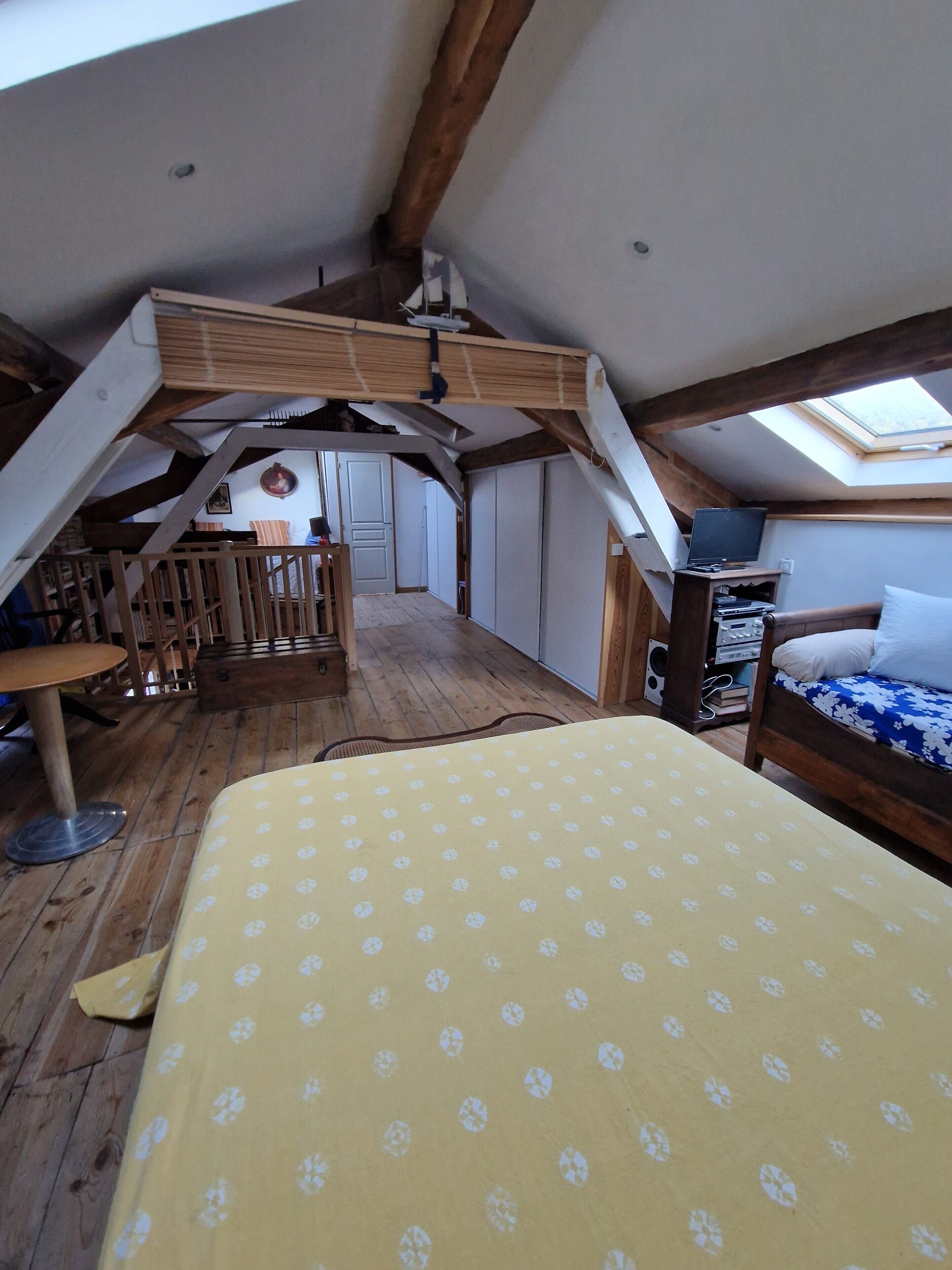 Une chambre à coucher aménagée dans un grenier, avec un plafond en pente, des poutres en bois, un lit recouvert d’une couvre-lit à motifs floraux jaunes, un bureau en bois et un téléviseur.