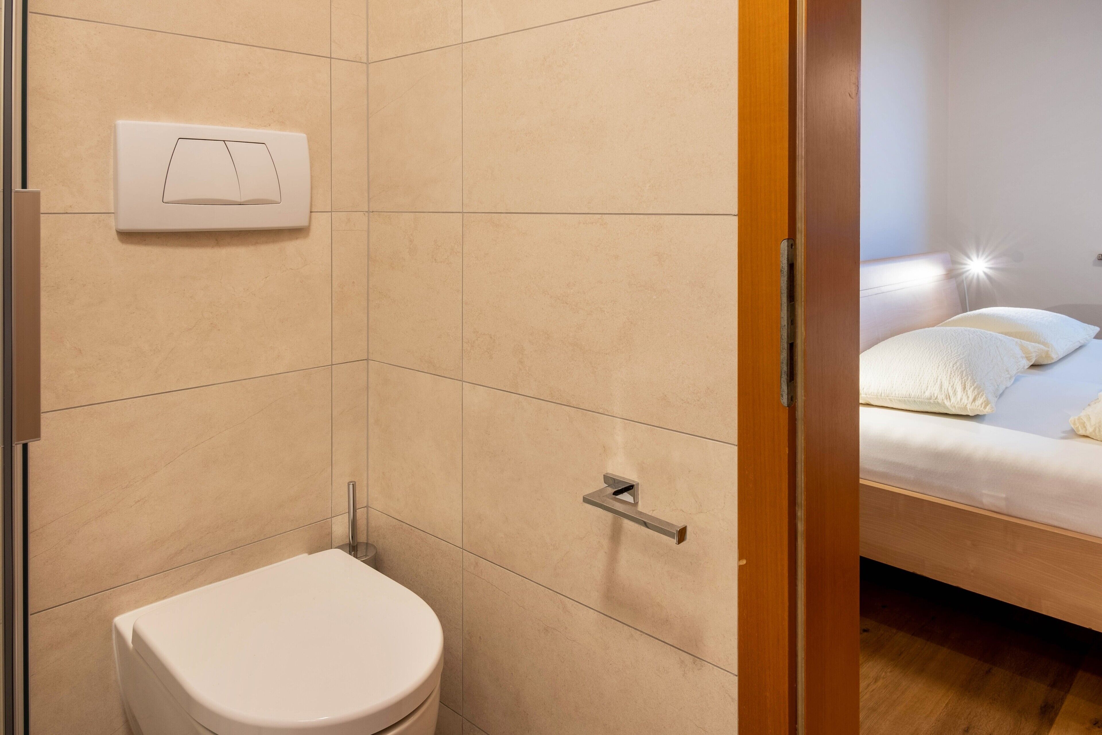 A bathroom with a toilet, a shower, and a bed visible through an open door.