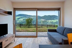 A modern living room with a large sliding glass door leading to a balcony with a scenic view of rolling hills and a wooden fence.