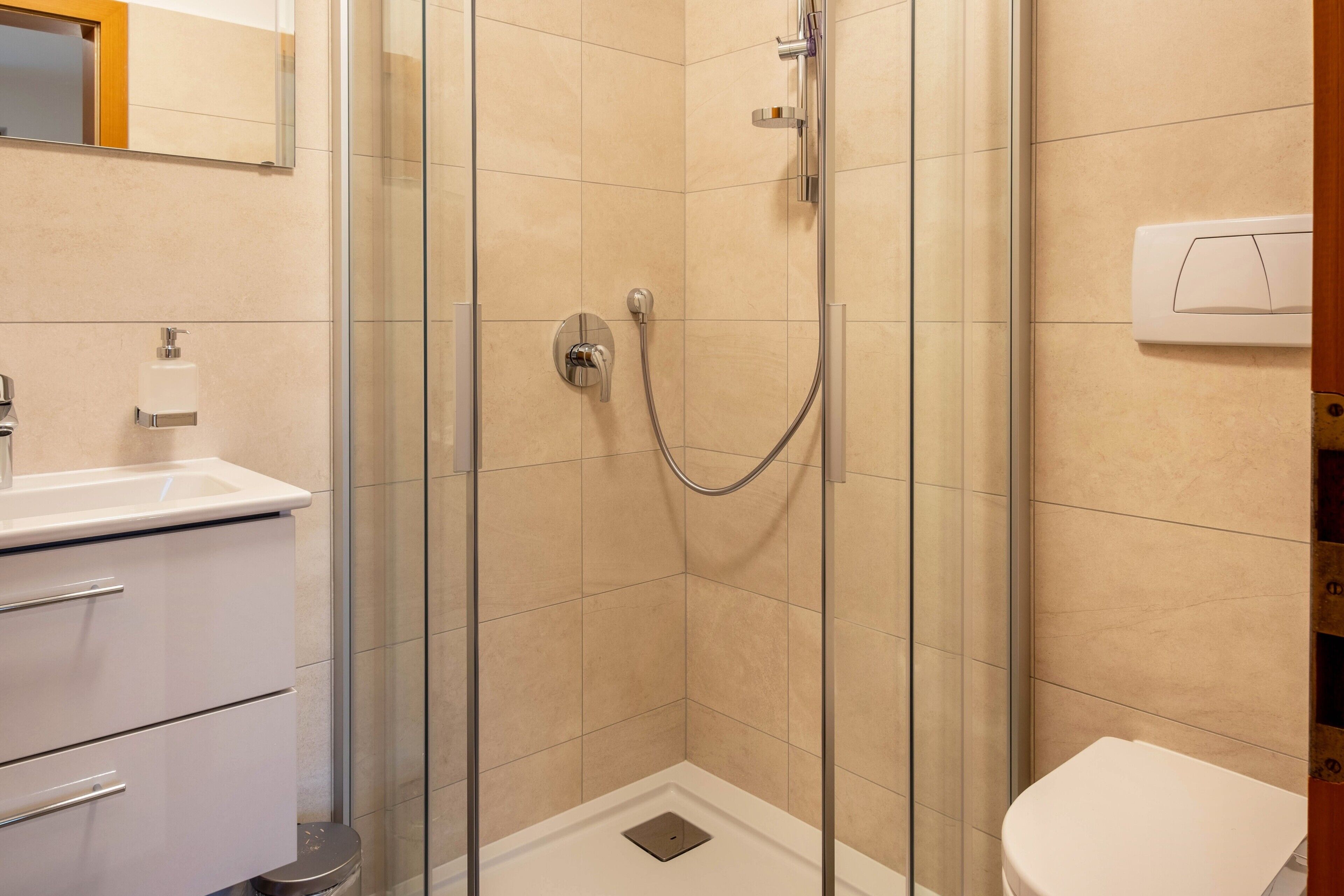 A bathroom with a glass-enclosed shower, a toilet, a sink with a soap dispenser, and a mirror.