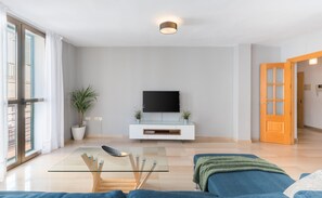 A modern living room with a glass coffee table, a flat-screen TV mounted on a white cabinet, a large window with curtains, and a wooden door.