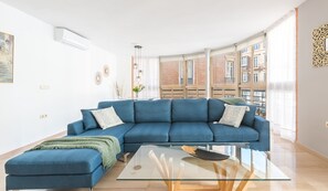 A modern living room with a blue sofa, a glass coffee table, and large windows offering a view of buildings.