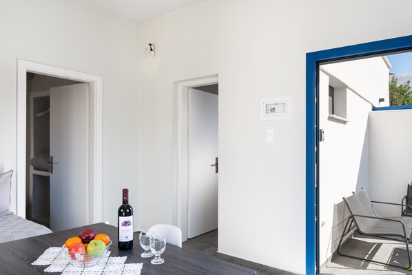 A room with a dining table set with fruit, wine, and glasses, a bed with a pillow, and a view of the outside. - Aegean Blue Santorini Perissa II near the beach with Private Terrace & Wi-Fi (Santorini)