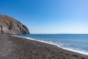A beach with dark sand, a rocky cliff, and calm blue water. - Aegean Blue Santorini Perissa II near the beach with Private Terrace & Wi-Fi (Santorini)