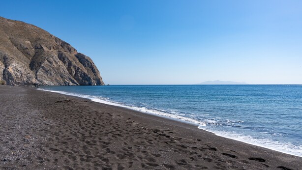 Ein Strand mit dunklem Sand, einer felsigen Klippe und ruhigem, blauem Wasser.