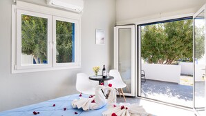 Ein Zimmer mit einem Bett, einem Tisch mit Flasche und Gläsern, weißen Stühlen und Blick auf Bäume durch eine Glastür.