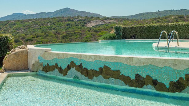 Ein Schwimmbecken mit Fliesenrand und Metallleiter, umgeben von Grünflächen und Hügeln im Hintergrund.