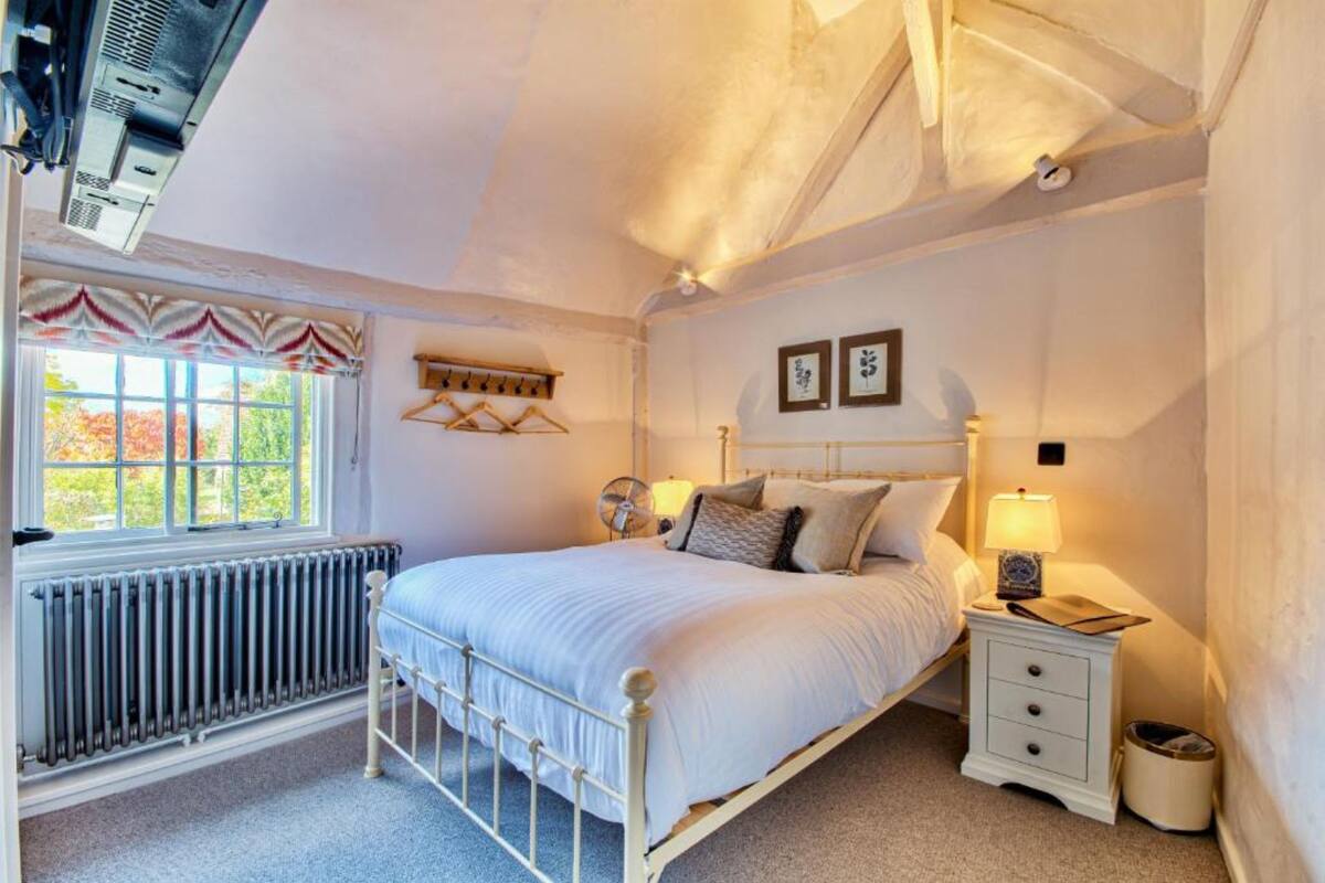 Une chambre à coucher comprenant un lit, une table de chevet, une lampe, un radiateur, une fenêtre avec des rideaux et des tableaux accrochés au mur.