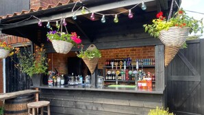 An outdoor bar with a rustic wooden counter, hanging flower baskets, and a variety of bottles and glasses. - The WoolPack Inn (Colchester)
