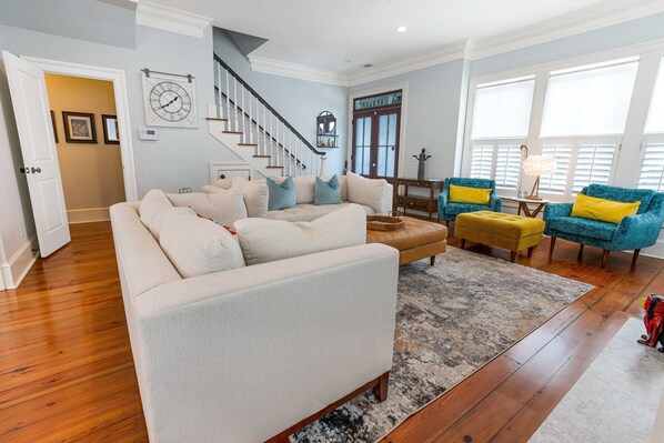 A spacious living room with a beige sofa, a blue armchair, and a wooden coffee table. A staircase is visible in the background, leading to another level. - Bright, Luxurious Executive Rental Near Forsyth w/ Parking by Lucky Savannah (Savannah)