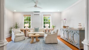 A spacious living room with a ceiling fan, two windows with views of greenery, a wooden cabinet, and a blue rug. - HONEY~ Adult Only, 2nd Floor 1 King Bedroom Unit w/ Community Pool! (Key West)