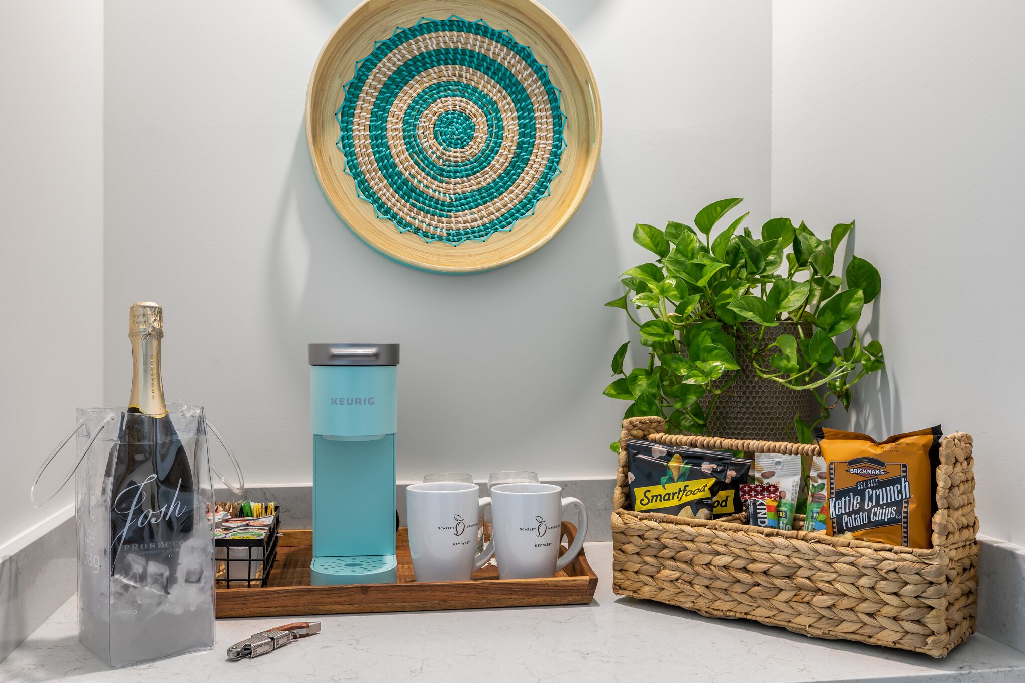 A kitchen counter with a bottle of champagne, a water dispenser, two mugs, a tray with food items, and a woven basket with snacks.
