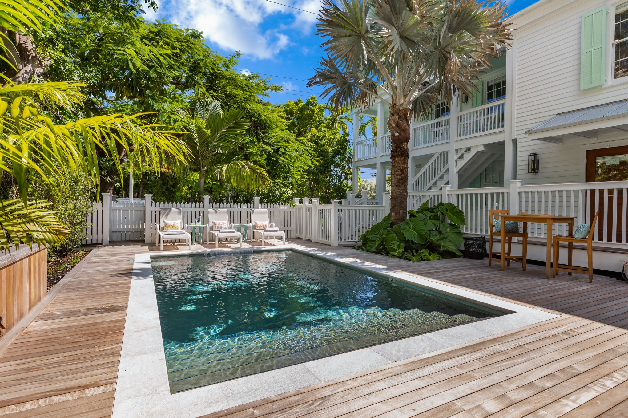 A swimming pool with a wooden deck, surrounded by lush greenery and a white fence.