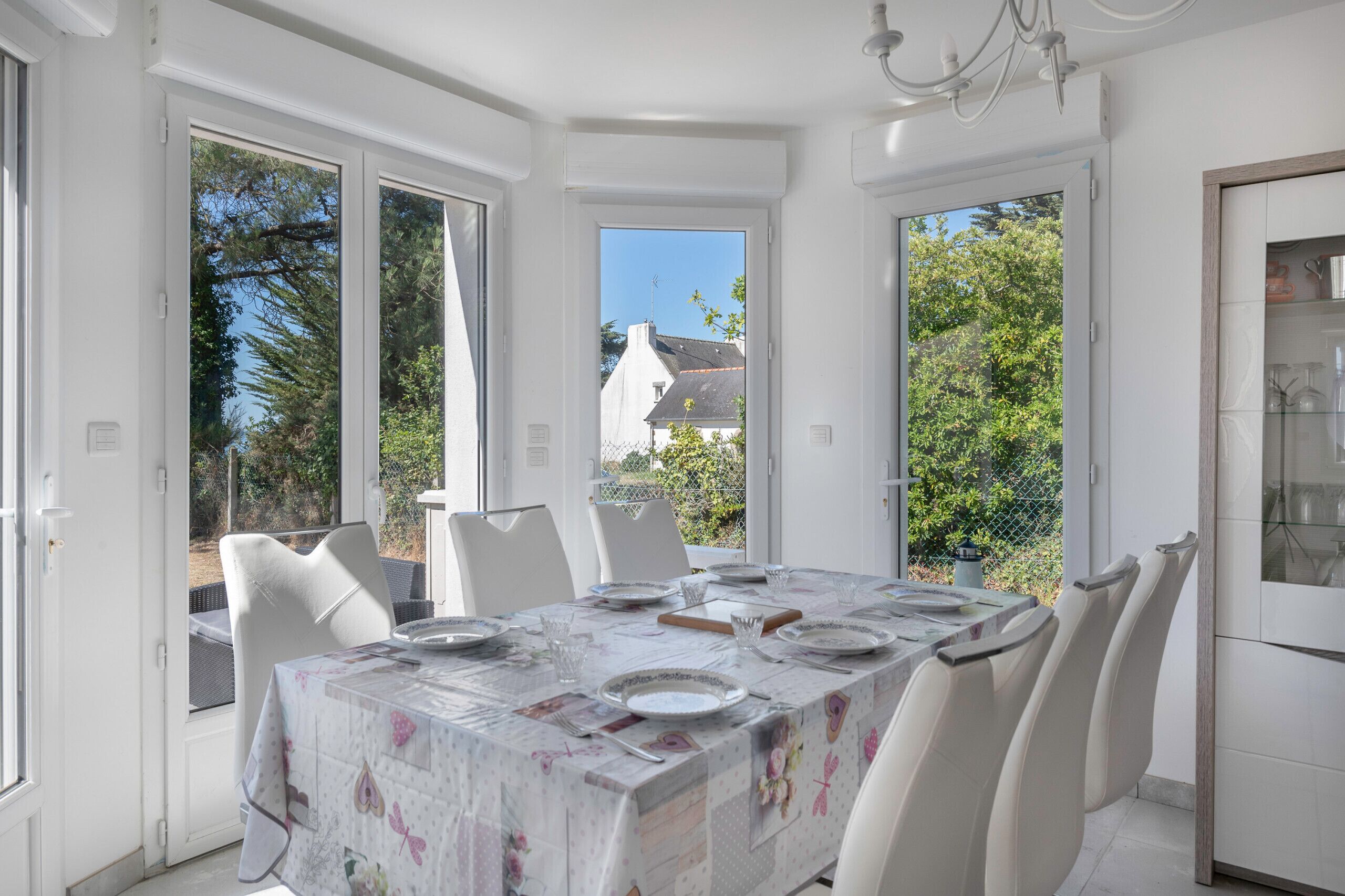 Un comedor con una mesa dispuesta para cuatro personas, sillas blancas y ventanas amplias que ofrecen vistas al exterior.