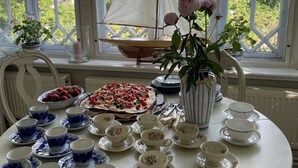 Ett matbord med ett urval av rätter och teserviser, en modell av en segelbåt och en vas med blommor, placerat framför stora fönster med utsikt över grönskan.