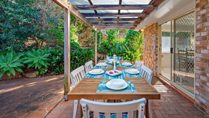 Eine Terrasse im Freien mit einem Holztisch, gedeckt für ein Essen, umgeben von üppigem Grün und einer Backsteinmauer.